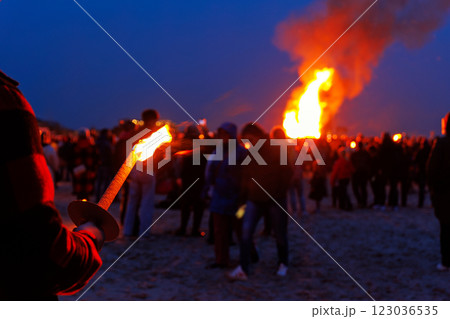 Scenic view of person hand with flaming torch against people crowd gathering celebrate traditional big Easter bonfire at twilight night in Germany. Walpurgis night fire party outdoors 123036535