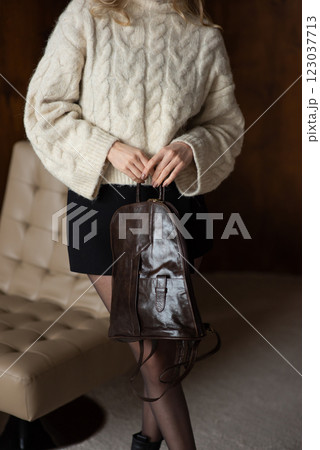 A girl in a warm sweater, a black short skirt and nylon tights is posing with a brown backpack. 123037713
