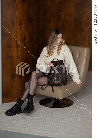 Woman in a warm sweater, a black short skirt and nylon tights is posing with a brown backpack. Woman in a warm sweater, a black short skirt and nylon tights is posing with a brown backpack. 123037745