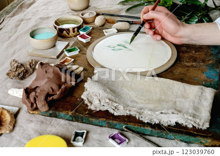 Workspace in ceramic studio, paints and brushes for underglaze painting. Young man's hands painting white plate. Branches, natural decor, linen tablecloth, daylight. Workshop idea. 123039760