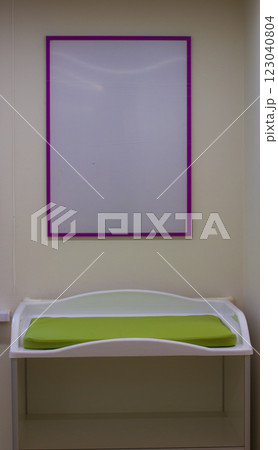 White baby changing table with green mattress and empty purple frame hanging on the wall in a nursery, representing childcare and baby care facilities. White baby changing table with green mattress and empty purple frame hanging on the wall in a nursery, representing childcare and baby care facilities. 123040804