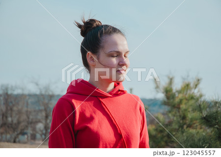 Young woman meditating outdoors in red hoodie on a sunny day 123045557