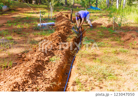 Plumber works diligently to dig trench install an PVC water pipe irrigation line in lush garden surrounded by trees. 123047050