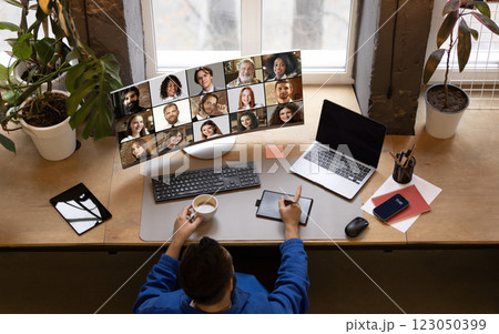 Overhead view of remote worker in blue jacket holding coffee, engaging in online meeting with large virtual team displayed on monitor against cozy home office. 123050399