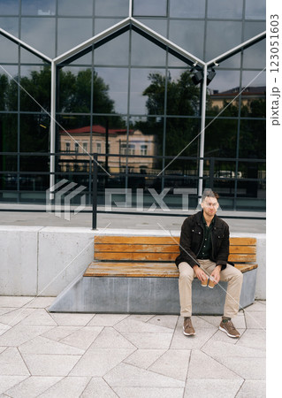 Full length vertical portrait of thoughtful man sitting on modern bench in city street, holding two disposable coffee cups, enjoying break in front of modern building. Concept of urban lifestyle. 123051603