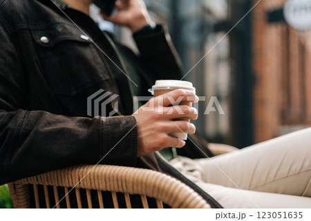 Cropped shot of unrecognizable man sitting o city a bench, enjoying moment of relaxation while sipping takeaway coffee and engaging in lively phone conversation, surrounded by hustle of urban life. Cropped shot of unrecognizable man sitting o city a bench, enjoying moment of relaxation while sipping takeaway coffee and engaging in lively phone conversation, surrounded by hustle of urban life. 123051635