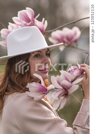 Woman magnolia flowers, surrounded by blossoming trees., hair down, white hat, wearing a light coat. Captured during spring, showcasing natural beauty and seasonal change. 123052110