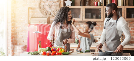Little black girl feeding mom while cooking together with parents at home, panorama 123053069