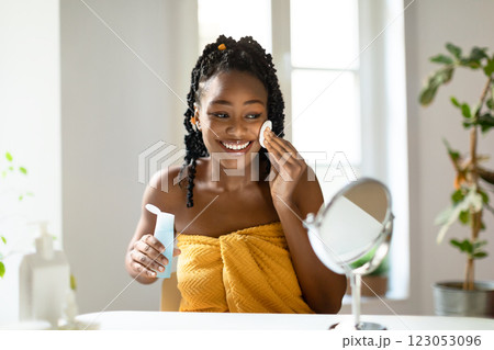 A black woman with dark braided hair, wearing a yellow towel, sits in front of a mirror and applies toner to her face with a cotton pad. 123053096