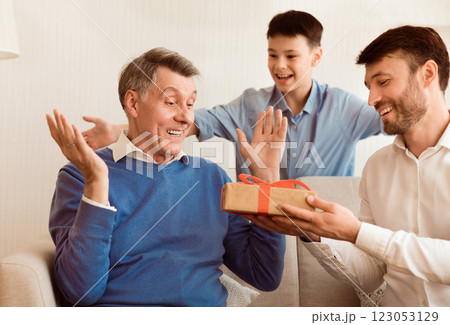 Middle-Aged Son And Grandson Congratulating Elderly Man Giving Him Birthday Gift Sitting On Couch Indoor. Selective Focus Middle-Aged Son And Grandson Congratulating Elderly Man Giving Him Birthday Gift Sitting On Couch Indoor. Selective Focus 123053129