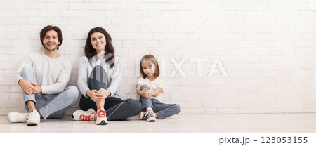Portrait of happy young parents and their cute little daughter sitting on floor in similar pose, leaning on white brick wall, empty space 123053155