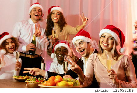 Joyful Multiethnic Friends In Santa Hats Holding Champagne Glasses Gesturing Victory Sign Smiling At Camera Celebrating New Year At Home 123053192