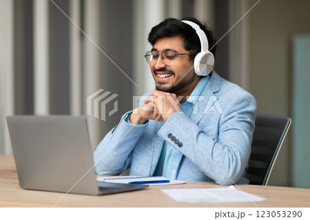 Indian man wearing headphones and glasses sits at a desk and smiles while looking at a laptop screen. He is likely participating in a video call with someone else. A notebook and pen are on the desk Indian man wearing headphones and glasses sits at a desk and smiles while looking at a laptop screen. He is likely participating in a video call with someone else. A notebook and pen are on the desk 123053290