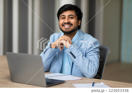 A young Indian man in a light blue blazer sits at a desk in a modern office. He is looking directly at the camera and smiling. His hands are clasped in front of him, and he has a laptop open 123053336