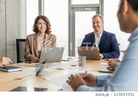 Three business professionals are sitting around a table in a modern office, using laptops and working on projects. Papers and writing utensils are spread out on the table 123053352