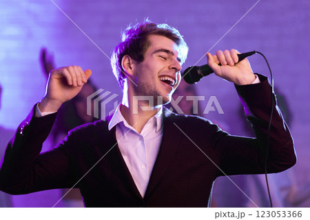Millennial Guy Singing Song Holding Microphone At Karaoke Night Party Indoor. Low Light Millennial Guy Singing Song Holding Microphone At Karaoke Night Party Indoor. Low Light 123053366
