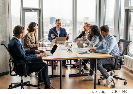 A group of five professionals are seated around a large, rectangular table in a modern office. They are engaged in a discussion, looking at each other and using laptops 123053402