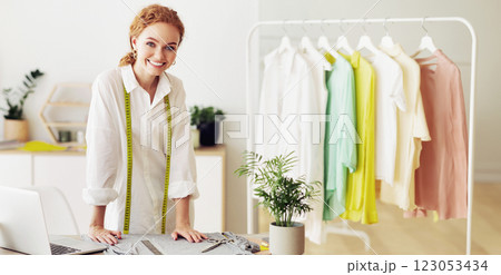 A woman with red hair, wearing a white shirt and a measuring tape, smiles while standing in a studio, with clothes hanging on a rack in the background. 123053434