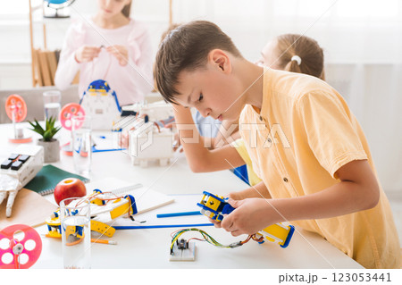 A young boy is focused on assembling a robot during a robotics class. He is sitting at a table with other students, all working on their own projects. 123053441