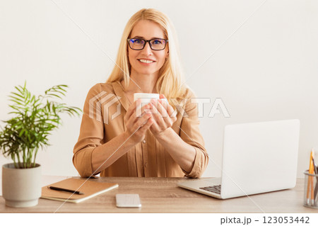 A woman sits at a desk, holding a coffee cup in her hands, with a laptop open in front of her. 123053442