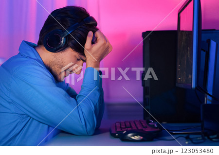 A man in a blue shirt sits at a desk with his head in his hands, looking disappointed. Hes wearing headphones and appears to be frustrated after losing a video game on a computer nearby. A man in a blue shirt sits at a desk with his head in his hands, looking disappointed. Hes wearing headphones and appears to be frustrated after losing a video game on a computer nearby. 123053480