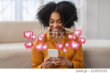 A young black woman with curly hair is sitting at home and smiling while checking her smartphone. She is surrounded by pink heart icons, indicating that she is receiving many likes 123053555