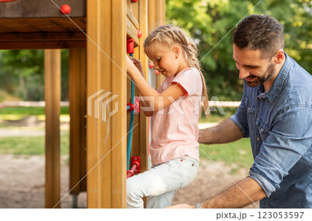 A father stands on the ground, encouraging his daughter who is climbing a rope ladder on a wooden playground structure. He is smiling and watching her as she works her way up the ladder. A father stands on the ground, encouraging his daughter who is climbing a rope ladder on a wooden playground structure. He is smiling and watching her as she works her way up the ladder. 123053597