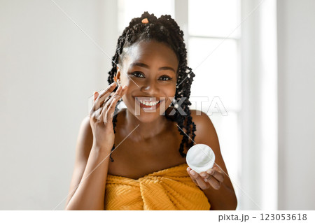 African American woman with braided hair smiles as she applies moisturizer to her face in a bathroom. She is wearing a yellow towel and is holding a container of moisturizer in her other hand. 123053618