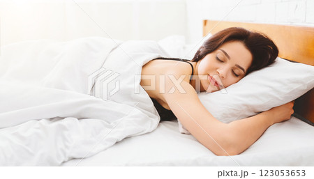 A woman is lying down in bed with her eyes shut, showing a moment of rest and relaxation. The room is dimly lit, and she appears calm and peaceful, panorama 123053653