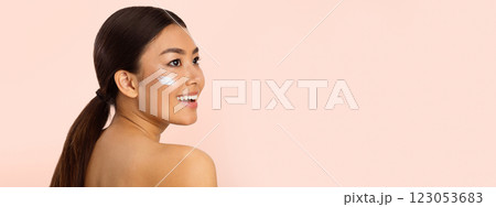 Asian young woman with long dark hair pulled back in a ponytail. She is smiling and looking to copy space with a dab of moisturizer on her cheek. The background is a soft pink color, panorama 123053683