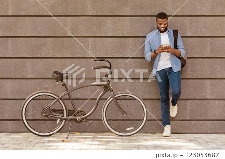 Young Black Man Using Smartphone, Standing Next To His Bike, Leaning On Grey Urban Wall, Free Space 123053687