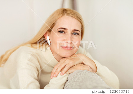 A woman with blonde hair is wearing earbuds and resting her head on her hands while sitting on a couch. 123053713