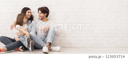 Casual Family. Portrait of cheerful mom, dad and their little daughter ebracing together and sitting near white brick wall on floor, copy space Casual Family. Portrait of cheerful mom, dad and their little daughter ebracing together and sitting near white brick wall on floor, copy space 123053714