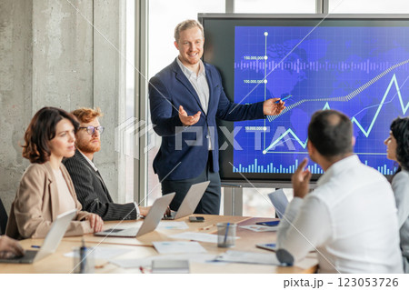 A confident businessman in a blue suit stands in front of a large screen displaying a blue and white graph. The graph shows a strong upward trend with a rising line, suggesting positive growth A confident businessman in a blue suit stands in front of a large screen displaying a blue and white graph. The graph shows a strong upward trend with a rising line, suggesting positive growth 123053726