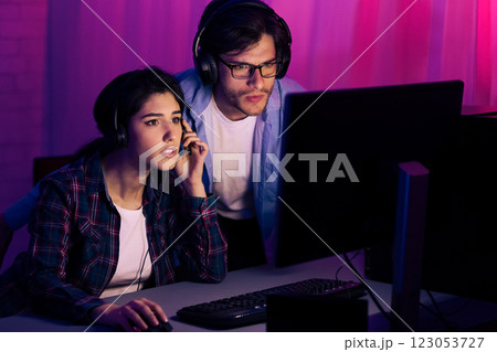 A young couple, both wearing headphones, sit in front of a computer screen in a dimly lit room. The man is standing behind the woman, both looking intently at the screen, likely engaged in a game. A young couple, both wearing headphones, sit in front of a computer screen in a dimly lit room. The man is standing behind the woman, both looking intently at the screen, likely engaged in a game. 123053727