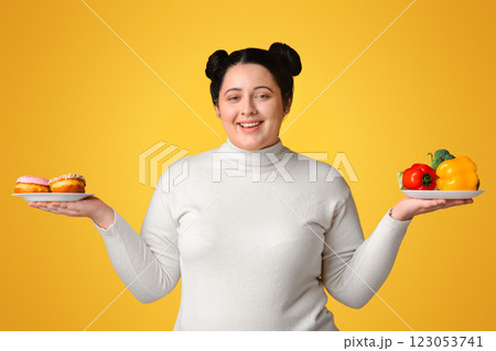 Smiling Plus Sized Girl Holding Plates With Fresh Vegetables And Donuts In Both Hands, Posing Over Yellow Background, Panorama 123053741
