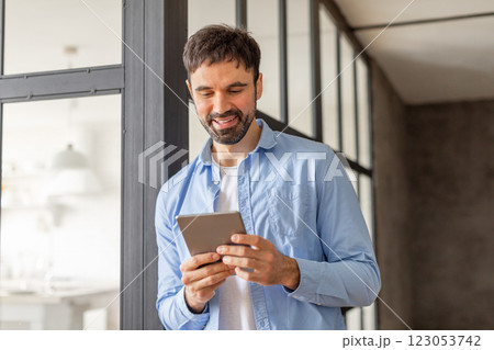 A man in a blue shirt stands in an office building, using a tablet device while looking at the screen with a smile. The man is in a casual work environment, surrounded by large windows. A man in a blue shirt stands in an office building, using a tablet device while looking at the screen with a smile. The man is in a casual work environment, surrounded by large windows. 123053742