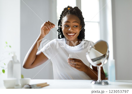 A young black woman with dark hair pulled back in braids smiles at her reflection while applying facial serum with a dropper. She is sitting at a vanity table with a small mirror A young black woman with dark hair pulled back in braids smiles at her reflection while applying facial serum with a dropper. She is sitting at a vanity table with a small mirror 123053797