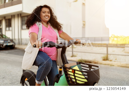 A young woman rides a bicycle down a city street wearing casual clothes, with a joyful expression on her face. The sun is shining, creating a lively atmosphere. A young woman rides a bicycle down a city street wearing casual clothes, with a joyful expression on her face. The sun is shining, creating a lively atmosphere. 123054149