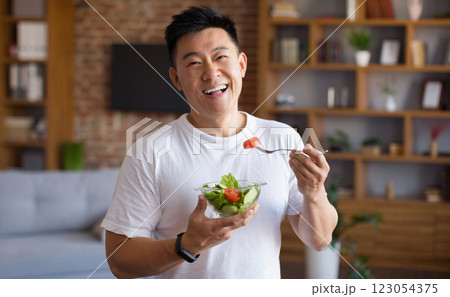 Happy asian mature man holding fork and bowl with fresh vegetable salad, eating healthy lunch after domestic training. Active middle aged male having balanced meal 123054375