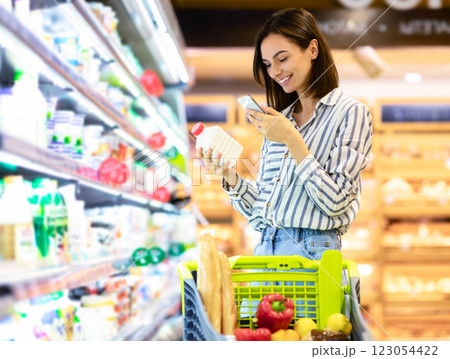 Smiling young woman taking dairy products from shelf in the supermarket, holding bottle and smartphone, scanning bar code on product through mobile phone, walking with trolley cart Smiling young woman taking dairy products from shelf in the supermarket, holding bottle and smartphone, scanning bar code on product through mobile phone, walking with trolley cart 123054422