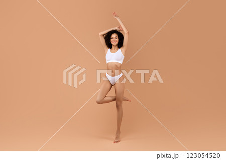 A young African American woman in white underwear poses in front of a tan background, smiling and looking at the camera. Her arm is raised above her head and she is balancing on one leg. 123054520