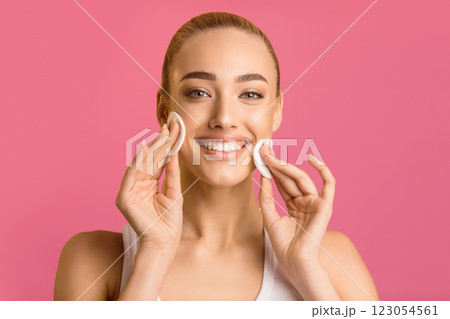 This image shows a smiling woman applying cotton rounds to her cheeks. She is wearing a white tank top and is standing in front of a pink background. Her face is clean and radiant This image shows a smiling woman applying cotton rounds to her cheeks. She is wearing a white tank top and is standing in front of a pink background. Her face is clean and radiant 123054561
