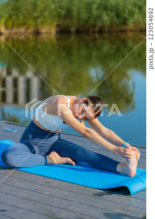 Fit Caucasian young woman in sportswear doing stretching exercising on yoga mat near pond in park 123054692