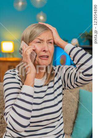Mature woman looking upset anxious while listening to bad news on mobile phone sitting on home sofa Mature woman looking upset anxious while listening to bad news on mobile phone sitting on home sofa 123054890