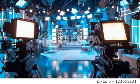 Two television cameras are positioned on tripods in a brightly lit television studio set. The set features a couch, two chairs, and a large screen. 123055131