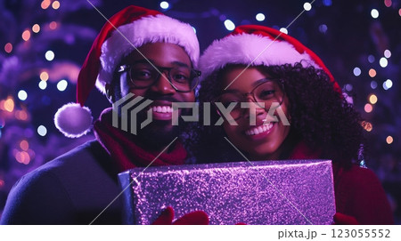 A couple in Santa hats stand in front of blurred Christmas lights, smiling at the camera. They are holding a silver gift box with their hands in front of their faces. 123055552