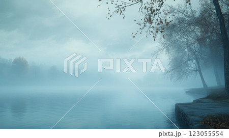 A calm lake reflects the soft hues of a misty morning, surrounded by autumn foliage. Trees border the shore, creating a serene atmosphere, with fog gently floating over the water's surface. 123055554