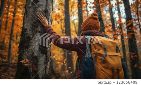 A person wearing a cozy beanie and carrying a backpack gently touches the trunk of a tree. Surrounding them are colorful autumn leaves, casting a warm glow in the fading sunlight. 123056409