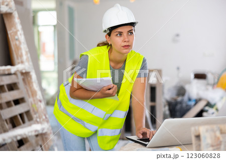 Female foreman in a protective helmet and a yellow vest checks the execution of repair work using laptop 123060828